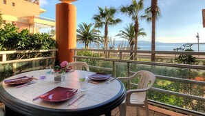 Mesa redonda con platos, vasos y un jarrón de flores en un balcón con vista a palmeras y al océano.