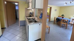 Een kleine keuken met een spoelbak, kookplaat en kastjes, die uitkomt op een eethoek met een tafel en stoelen.