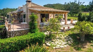 Une maison en pierre dotée d’un patio, entourée d’une végétation luxuriante et sous un ciel dégagé.