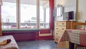 Una habitación con una cama, un aparador de madera, una mesa con mantel a cuadros, un televisor y ventanas grandes con vistas a edificios.