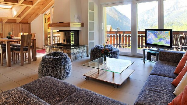 A spacious living room with a stone fireplace, a glass coffee table, and a large sofa. There is a dining area with wooden furniture and a view of mountains through large windows. - Edelweiss A by Interhome (Leukerbad)