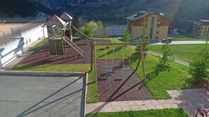 A playground with a slide and swings, surrounded by a building with a red roof and a mountain backdrop. - Edelweiss A by Interhome (Leukerbad)