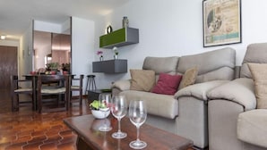 En stue med sofa, sofabord med vinglas, spiseplads med bord og stole, samt en vægmonteret hylde med pynt.
