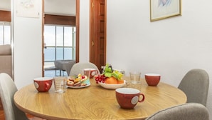 Et rundt træspisebord med fire stole, et morgenbord med frugt, kager og kaffekopper, og udsigt til havet gennem en åben dør.