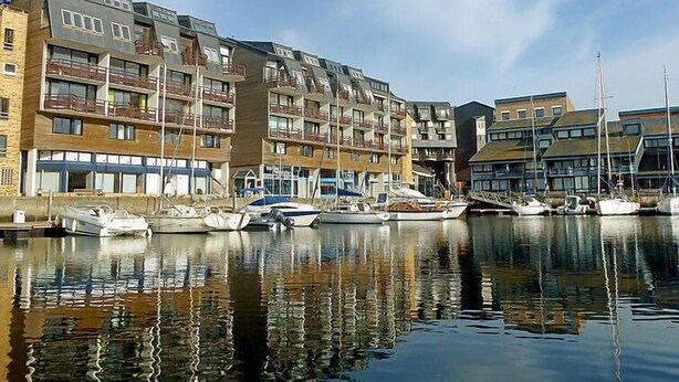 A marina with multi-story buildings, boats docked, and calm water reflecting the structures.