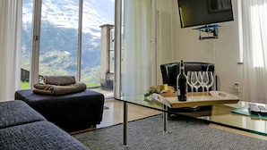 Un salon moderne avec une table en verre, un canapé, un téléviseur fixé au mur et une vue imprenable sur les montagnes, visible à travers de grandes fenêtres.