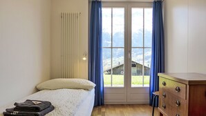 Une chambre à coucher comprenant un lit, une commode, un radiateur et une fenêtre donnant sur une grange et des montagnes.