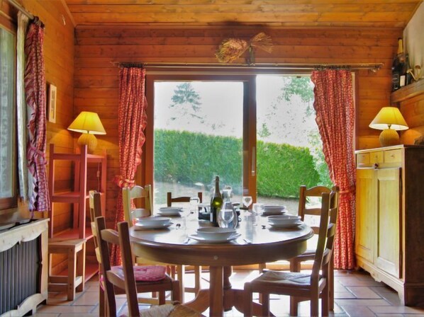 A wooden cabin dining room with a round table set for four, two lamps, and a view of a hedge outside.
