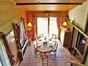 L’intérieur est en bois et comprend une table à manger pour six personnes, entourée de chaises. On y trouve deux fenêtres avec des rideaux, une cheminée et une étagère remplie d’objets, située à droite.