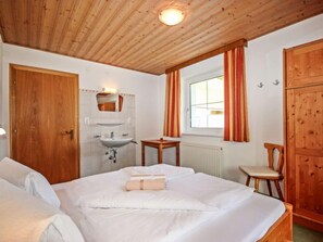 A bedroom with a wooden ceiling, a bed with white bedding, a wooden chair, a small table, a window with curtains, and a wooden wardrobe. - Apartment Sailer by Interhome (Kappl)