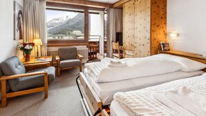 A hotel room with a bed, two chairs, a desk, a lamp, a window with a mountain view, and a wooden wardrobe. - Guardaval Apt 101 by Interhome (Davos)