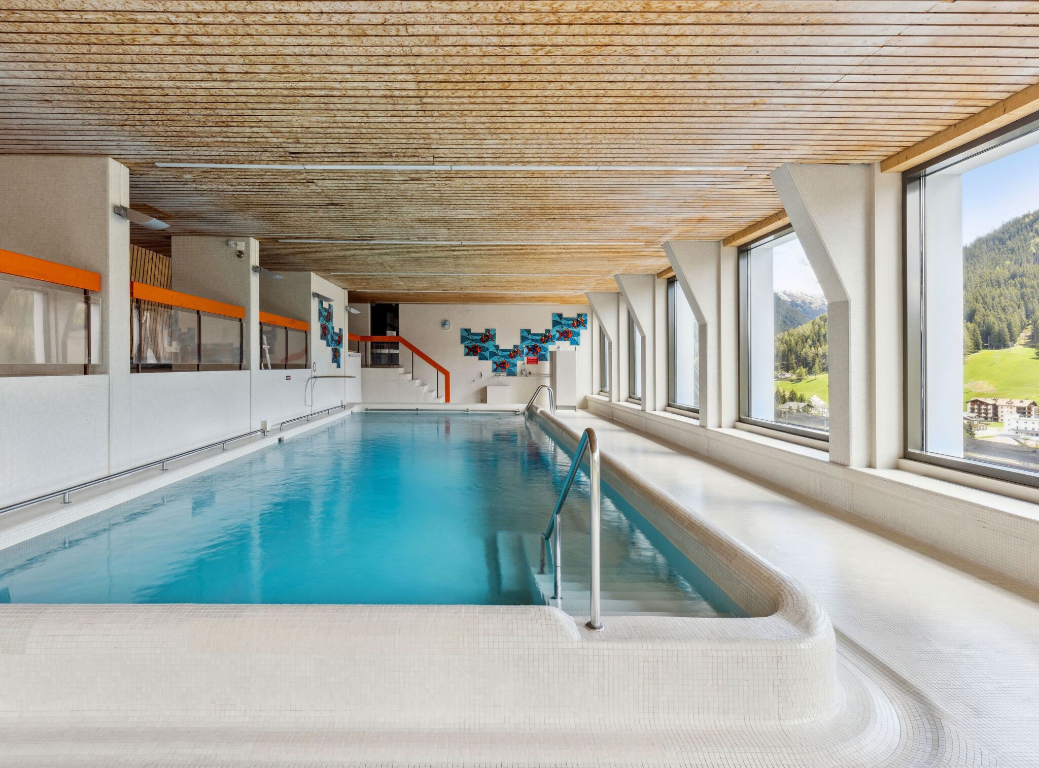 Una piscina interior con techo de madera, una escalera y vistas a un paisaje montañoso a través de amplios ventanales.