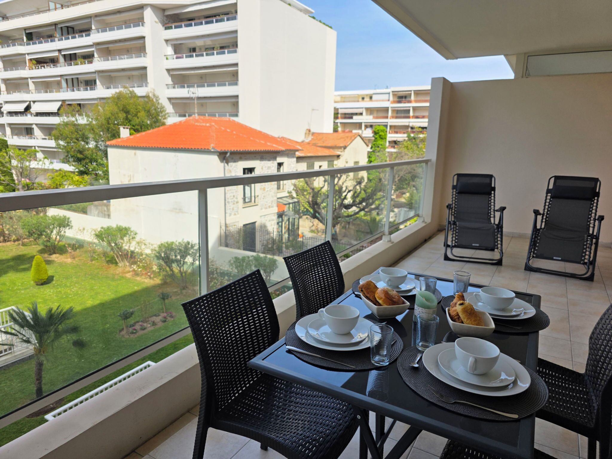 Un balcón con una mesa dispuesta para dos, sillas y vistas a un edificio y follaje.
