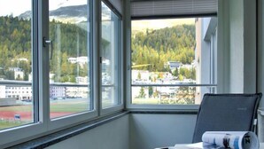 A room with a large window offering a view of a sports field and buildings. There is a chair with a rolled-up magazine on a table. - Chesa Ova Cotschna 205 by Interhome (St. Moritz)