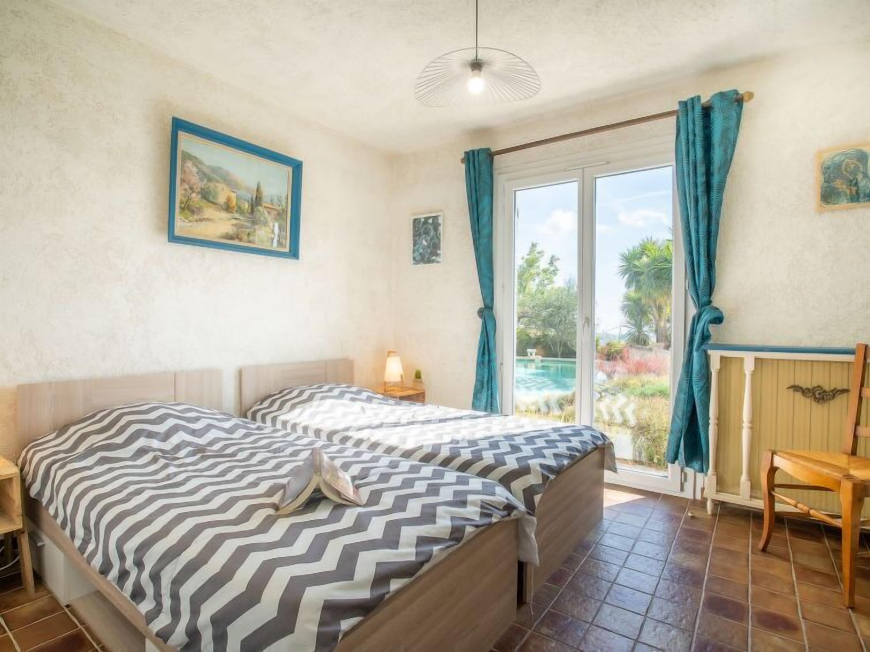 A bedroom with two beds, a window with curtains, a painting on the wall, and a view of the outside.