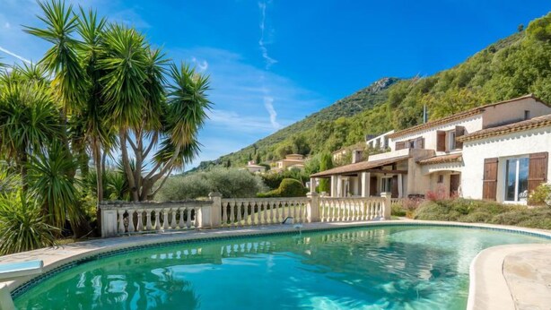 A swimming pool with clear water, surrounded by a white building with a tiled roof, and palm trees. - La Mesnière by Interhome (Vence)