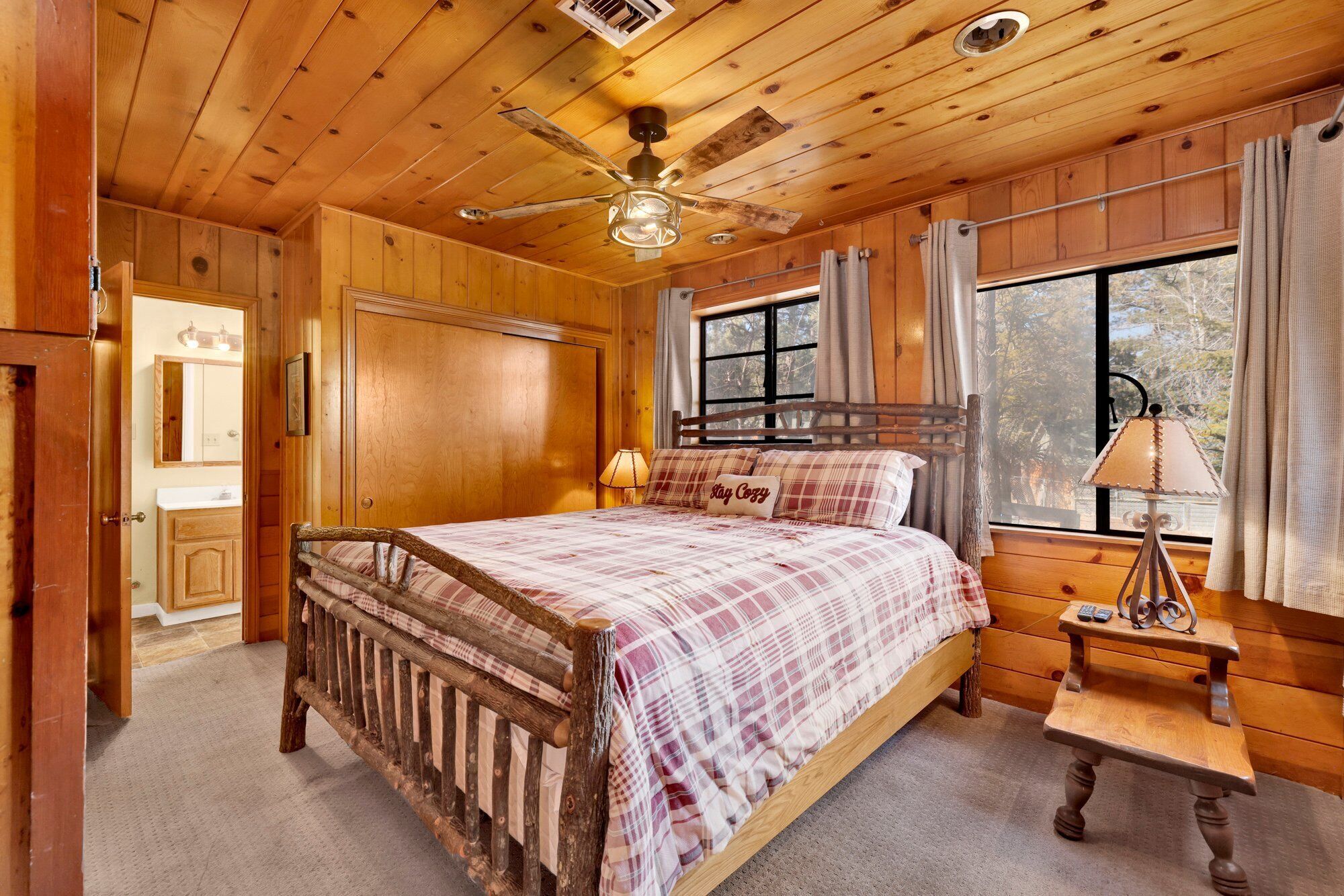 A wooden cabin bedroom with a bed, a nightstand, a ceiling fan, and a window with curtains.