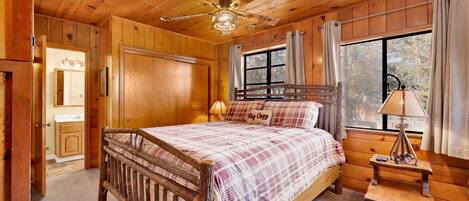 A wooden cabin bedroom with a bed, a nightstand, a ceiling fan, and a window with curtains.