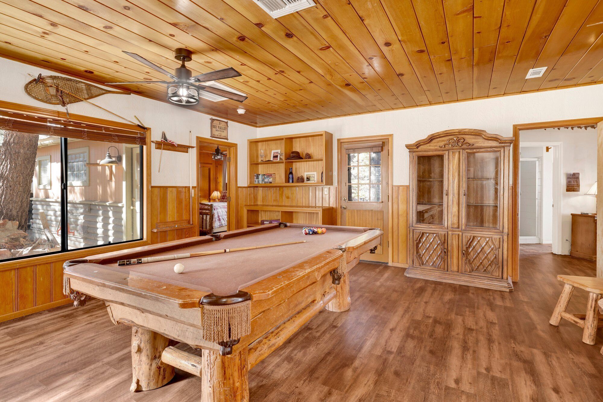 A pool table in a room with wooden walls and ceiling, a ceiling fan, and a wooden cabinet with glass doors.