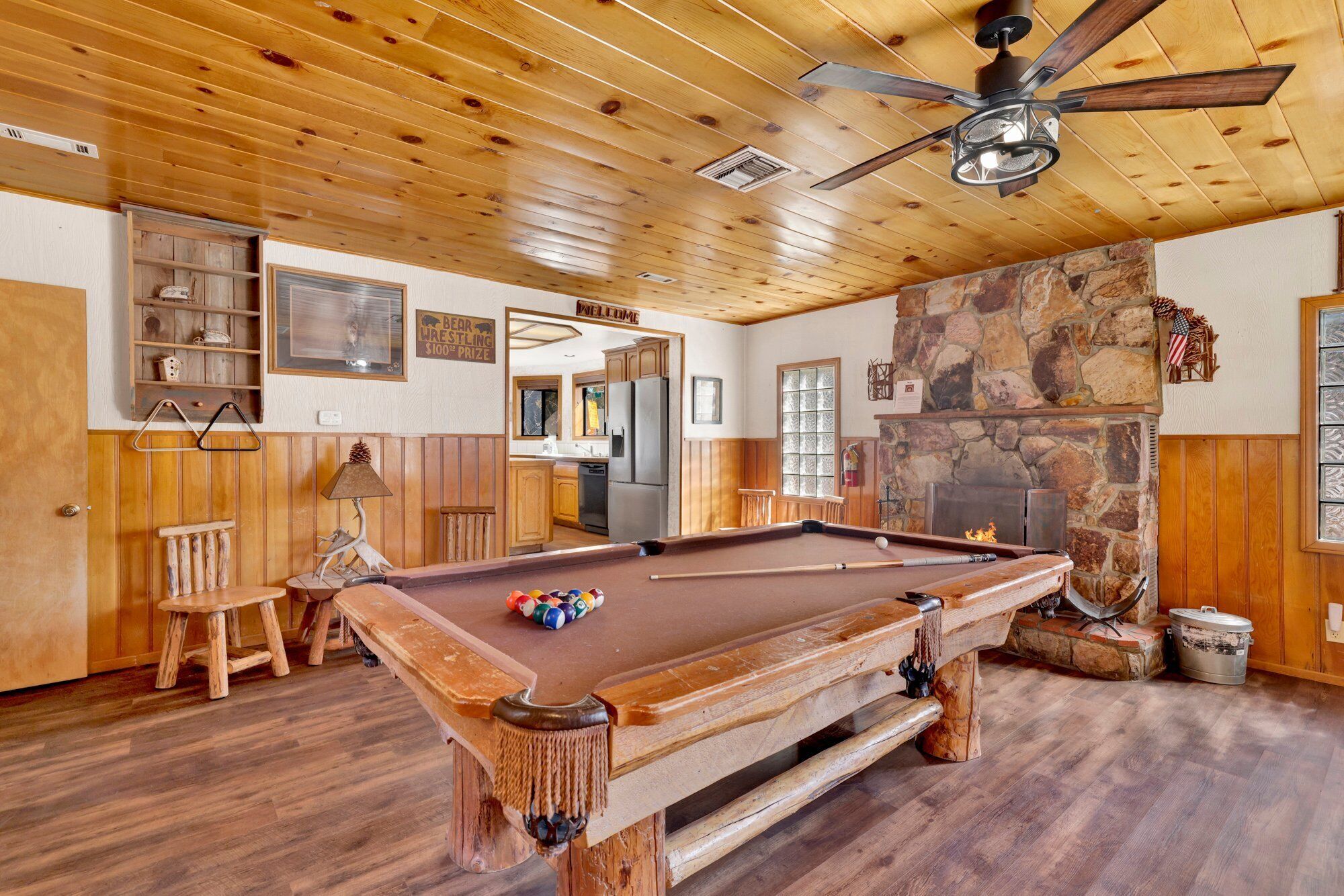 A wooden pool table in a room with a stone fireplace, wooden ceiling, and a ceiling fan.