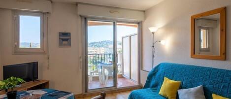 A living room with a sofa, a dining table set for two, a TV, and a balcony with a view.