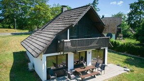 Una casa de dos pisos con un balcón, un patio con mobiliario exterior y un gran ventanal con vistas a un jardín extenso.