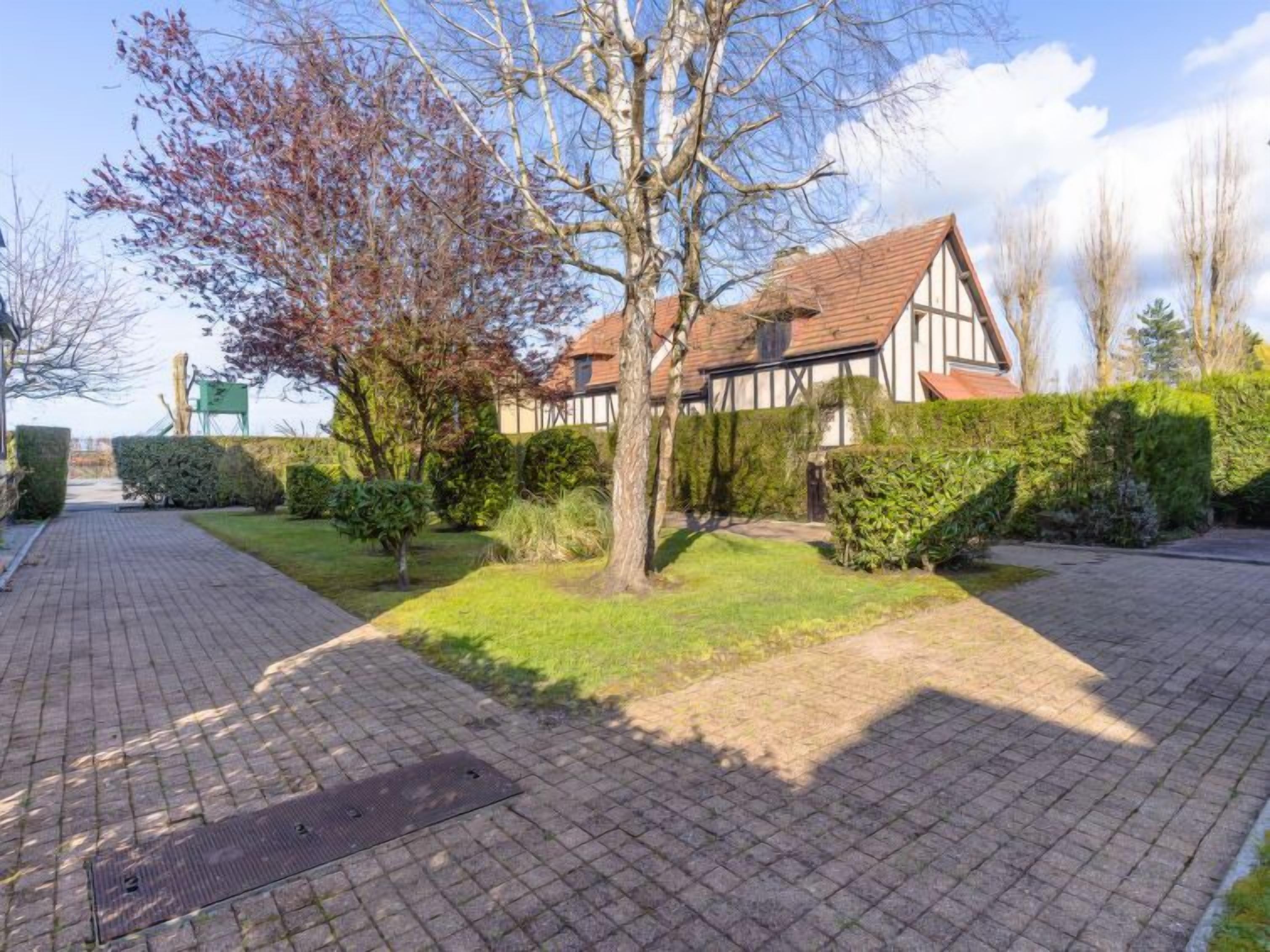 A traditional half-timbered house with a paved courtyard, surrounded by well-maintained hedges and trees.