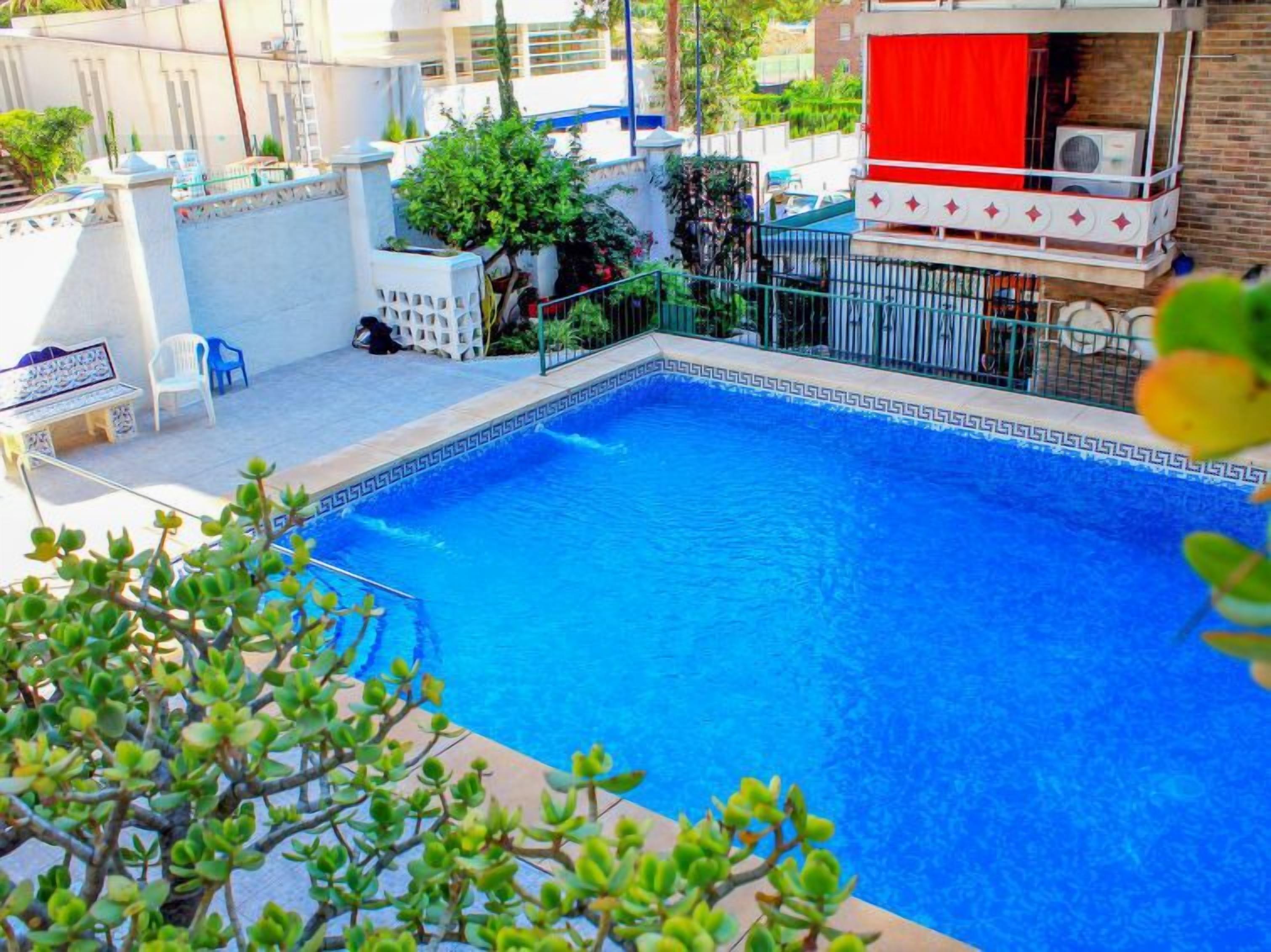 A swimming pool with a geometric tile border, surrounded by a white fence, and a red curtain on a balcony.