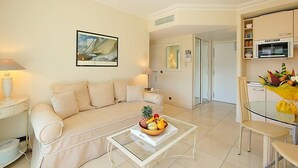 Ein Wohnzimmer mit einer Couch, einem Glastisch mit Obst, einem Gemälde und einer Küchenzeile mit einer Mikrowelle.