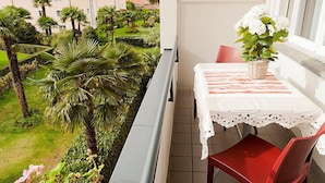 Un balcon avec une table et des chaises, donnant sur un jardin planté de palmiers et offrant une vue sur des maisons au loin.