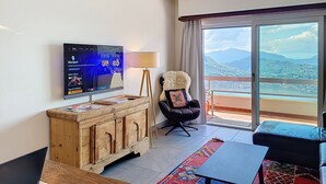 Habitación de hotel con televisor, aparador de madera, una silla negra, sofá y balcón con vista a montañas.