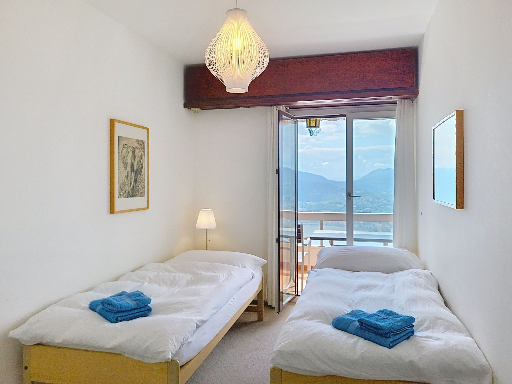 A hotel room with two beds, a large window with a view of mountains, a hanging light fixture, and a framed picture on the wall.