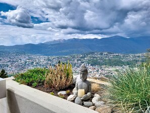 Una estatua de una figura sentada con una vela, rodeada de plantas y rocas, con vistas a un paisaje urbano y montañas al fondo.