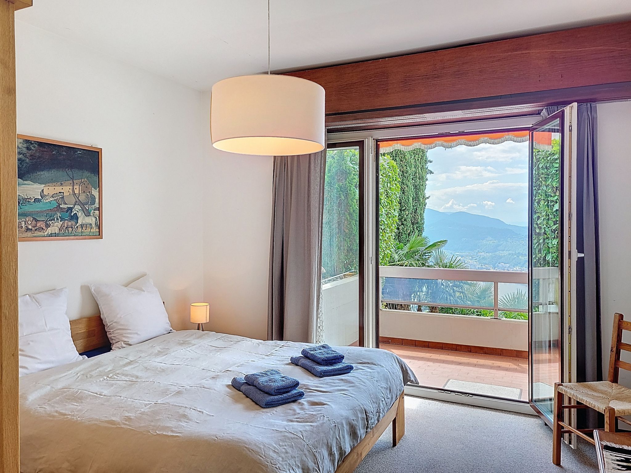 A bedroom with a bed, a painting, a lamp, and a view of greenery and mountains through a sliding door.