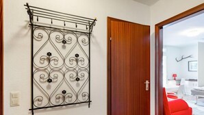 Ein Zimmer mit einer Holztür, einem Metallregal verziert mit Ornamenten, und ein Blick auf einen roten Stuhl sowie weiße Möbel im angrenzenden Raum.