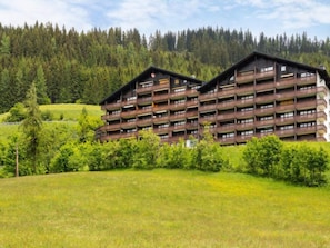 A multi-story building with balconies, surrounded by greenery and a forested hillside.