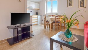 A modern living room with a flat-screen TV, a dining area with a table and chairs, a kitchen in the background, and a large window providing natural light.
