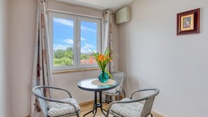 A room with a window, two chairs, a small round table with a vase of flowers, and a framed picture on the wall.