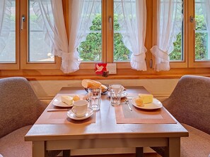 Une table de petit-déjeuner dressée avec des assiettes, des verres et un panier à pain, avec une vue sur la fenêtre et des rideaux.