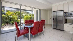 Une cuisine moderne équipée d’un réfrigérateur, d’un four à micro-ondes et d’un four, tous en acier inoxydable. On y trouve une table à manger avec des chaises rouges et un plateau en verre. La cuisine est dotée de grandes fenêtres qui offrent une vue sur un jardin et des arbres.