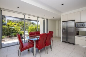 A modern kitchen with a stainless steel refrigerator, microwave, and oven. There is a dining table with red chairs and a glass top. The kitchen has large windows offering a view of a garden and trees.