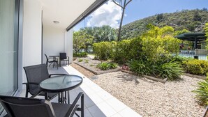 Un balcon avec une table et des chaises en verre, une vue sur un jardin luxuriant et une colline en arrière-plan.