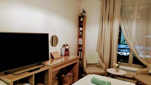 Ein Hotelzimmer mit Bett, Fernseher, Bücherregal, Sessel, kleinem Tisch und einem Fenster mit Vorhängen.
