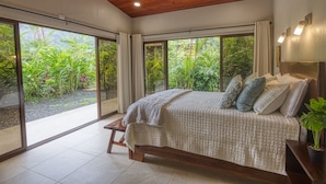 A bedroom with a large bed, wooden ceiling, and a view of lush greenery through large windows. - Sleeps 8, Brand New, private pool, steps from beach, dining and shopping (Dominical)