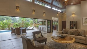 A spacious living area with a large window overlooking a pool and lush greenery. There is a seating area with a sofa and chairs, and a wooden coffee table. - Sleeps 8, Brand New, private pool, steps from beach, dining and shopping (Dominical)