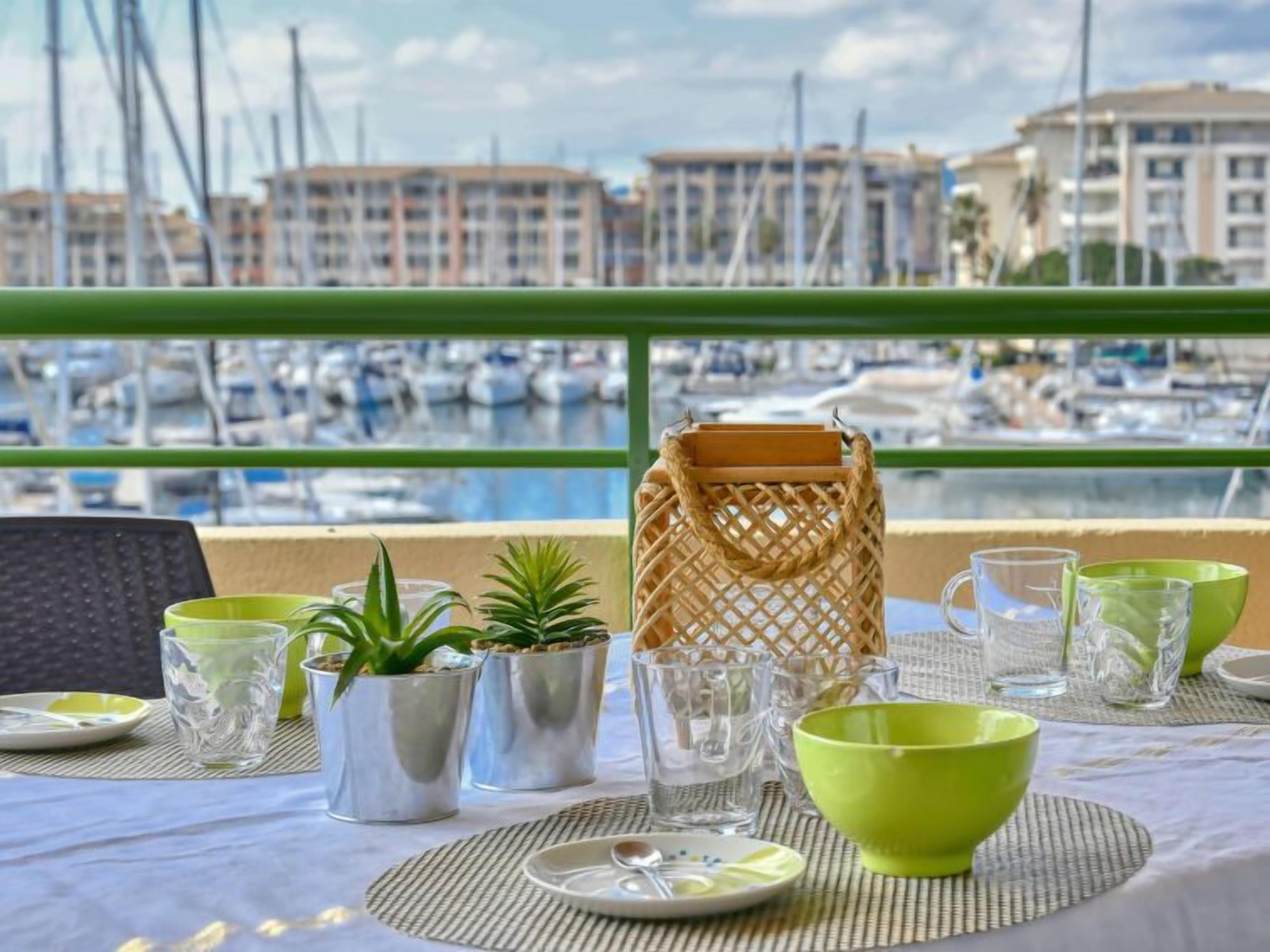 Une table dressée avec de la vaisselle transparente, des bols verts et des plantes en pot, donnant sur une marina où l’on peut admirer des bateaux et des bâtiments.