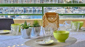 Et bord dækket med blankt glas, grønne skåle og potteplanter, med udsigt over en havn med både og bygninger.
