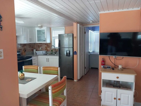 Cocina compacta con alacenas blancas, refrigerador de acero inoxidable, mesa de comedor con sillas a rayas y televisor sobre un mueble.