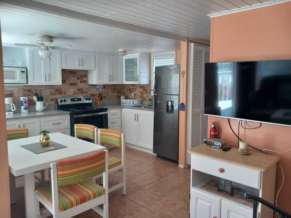 A compact kitchen with white cabinets, a stainless steel refrigerator, a stove, and a microwave. There is a dining table with striped chairs and a television on a stand.