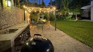 Un área de cocina al aire libre con una parrilla, un lavamanos de piedra y una pérgola con luces colgantes.